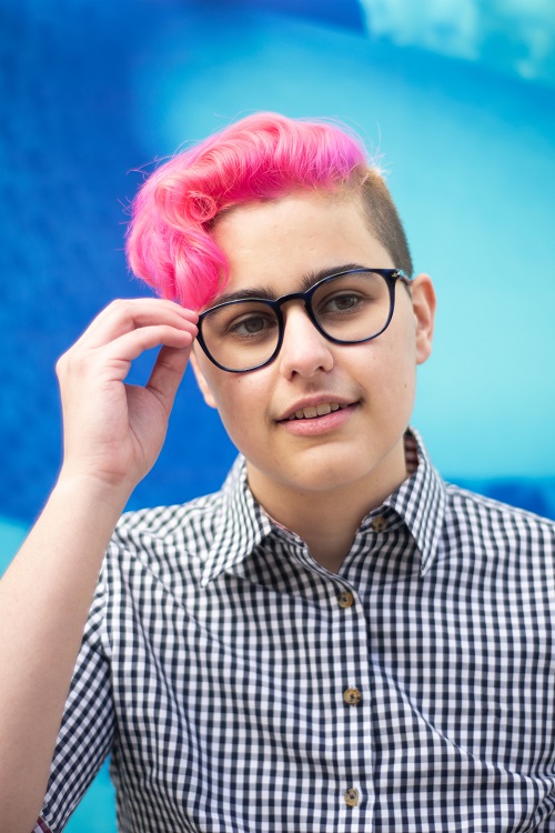 Lewis Figun Westbrook, a white nonbinary person with pink hair, touches their glasses with one hand. Behind them is a blue mural