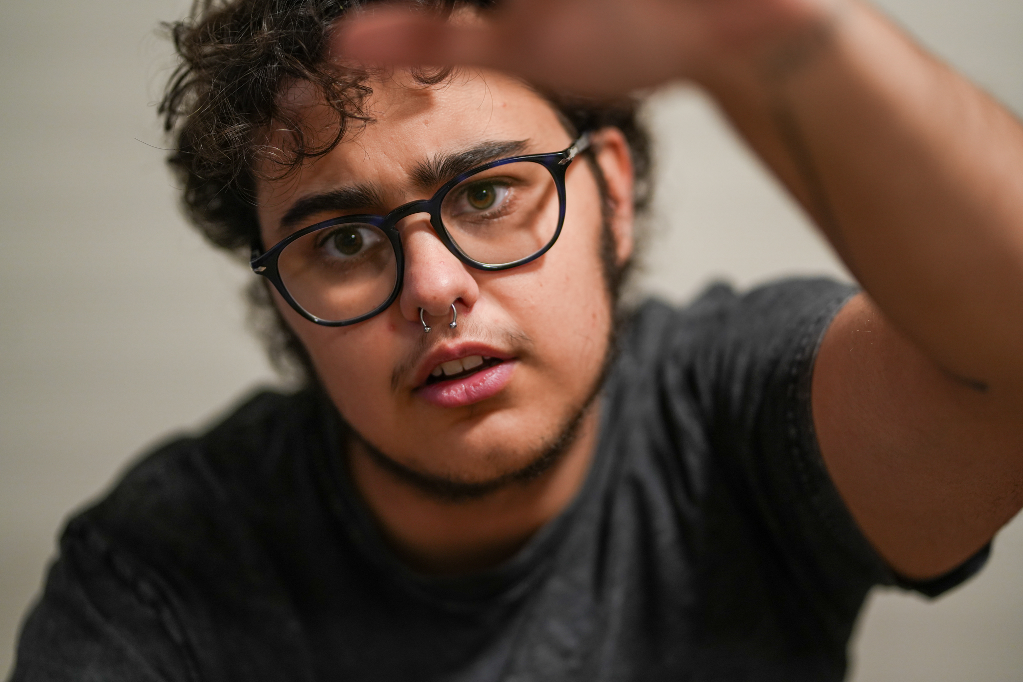 Lewis is a light skin queer person with curly brown hair and glasses. They have striking eyebrows, facial hair, and a septum nose piercing. They are wearing a grey shirt that is blurred and one hand is reaching out to the top of the frame. There are tattoos on their hand.